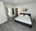Modern bedroom with a double bed, white bedding, nightstand, mirror, and artwork on the wall. The room features light-colored walls and a dark wooden floor.