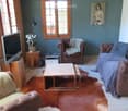 Living room with wooden ceiling, large windows, grey sofa, coffee table, armchairs, TV, and a painting on the wall.