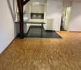 Empty room with wooden flooring, white walls, and a small kitchen area visible in the background. No furniture or furnishings are present.