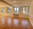 Empty room with wooden flooring, white walls, and multiple windows allowing natural light. Radiators are visible under the windows.