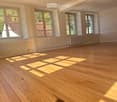 Empty room with wooden flooring, white walls, and large windows allowing natural light to enter. No furniture or equipment is present.