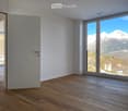 Empty room with wooden flooring, large windows offering a mountain view, and a closed door.