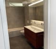Modern bathroom with a dark brown vanity, white sink, large mirror with built-in lighting, bathtub, and toilet. The walls are tiled in a neutral gray color, and the floor is covered with dark brown tiles.
