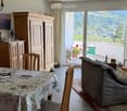 Bright living room with a view of the mountains, featuring a sofa, dining table with chairs, wooden cabinet, and large windows with curtains.