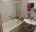 A small bathroom with a white tiled wall and floor, featuring a bathtub, a sink with a faucet, a showerhead, a towel on the floor, and a shelf with toiletries.