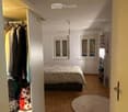 Cozy bedroom with wooden flooring, a bed with a patterned duvet, a wardrobe with hanging clothes, a bookshelf, and a round rug on the floor.