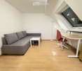 Modern, compact living space with a grey sectional sofa, small white coffee table, red office chair, white desk, and a floor lamp. The room features light wooden flooring and white walls, with a large window providing natural light.
