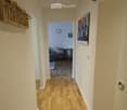 A narrow hallway with light wooden flooring, white walls, and recessed ceiling lights. A small shelf with decorative items is mounted on the left wall, and a painting hangs on the right wall. The hallway leads to an open living area with a sofa, chairs, a