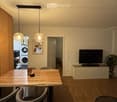 Bright, modern dining area with a wooden table, chairs, pendant lights, a TV on a cabinet, and a plant near the window. A private washing machine is visible in the adjacent room.
