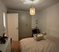 Cozy bedroom with a wooden floor, a large bed with white bedding, a wardrobe, a desk with a chair, a hanging woven lamp, and a small plant on a nightstand.