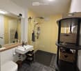 Modern bathroom with dark tiled floor, white sink, toilet, large mirror, and a glass-enclosed shower area with multiple showerheads. A black shelving unit holds various items, and towels are neatly arranged.