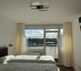 Bright bedroom with two single beds, large window with balcony access, and a wall-mounted TV.