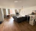 A modern, open-plan living and dining area with wooden flooring, large windows, a grey sectional sofa, a coffee table, a dining table with chairs, and a TV stand.
