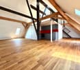 Open-plan attic space with exposed wooden beams, hardwood flooring, and a small kitchen area with dark cabinets and a red backsplash.