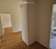 Empty hallway leading to a bathroom with a bathtub, toilet, and tiled walls. The hallway has wooden flooring and white walls.