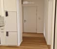 Empty hallway with wooden flooring, white walls, and a closed door at the end. A white cabinet and a refrigerator are visible on the left side.