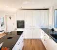Modern kitchen and living area with white cabinets, black countertops, a large window with a scenic view, a sofa, a dining table, and various kitchen appliances.
