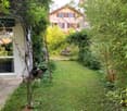 A lush garden with a grassy lawn, various plants, a decorative metal sculpture, a bench, and a pathway leading to a house entrance.