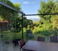 Garden with a wooden table and chairs, sun lounger, swing, fire pit, and potted plant, surrounded by lush greenery and a clear view of mountains.