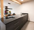 Modern kitchen with dark grey cabinets, a central island, and a dining area with a wooden table and chairs. The kitchen features a sleek sink and faucet, and there are vases with flowers on the table.