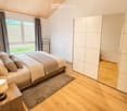 Cozy bedroom with a large bed, nightstand with lamp, and a modern wardrobe. The room features wooden flooring and large windows with curtains, offering a view of a green landscape.