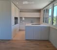 Modern kitchen with white cabinets, central island, and large windows offering a view of greenery.
