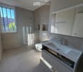 Modern bathroom with a large window, white sink, toilet, mirrored cabinet, and a glass-enclosed shower area.