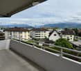 Balcony with white railing, overlooking a residential area with houses, trees, and distant mountains.