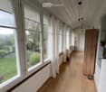 Bright hallway with wooden flooring, large windows with white curtains, and a view of a green garden outside.