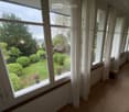 Bright hallway with large windows and white curtains, leading to a view of a well-maintained garden with shrubs and a small shed.