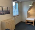 A small bedroom with a single bed, a wooden cabinet, a radiator, and three framed mirrors on the wall. The room has a window with white curtains and a pendant light fixture.