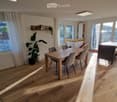 Bright, open-plan living and dining area with wooden flooring, large windows, and modern furniture including a dining table, chairs, and a potted plant.