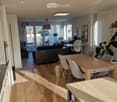 Open-plan living and dining area with wooden flooring, large windows, and a balcony. Features a sofa, dining table with chairs, and decorative plants.