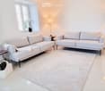 Modern living room with white walls, light-colored sofas, a white rug, and large windows letting in natural light. Includes a side table, floor lamp, and decorative vases.