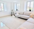 Modern living room with white walls, large windows, a beige sofa, a white chair, a small table with a laptop, a floor lamp, and decorative vases on the windowsills.