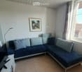 A modern living room with a blue sectional sofa, decorative pillows, a white side table, a floor lamp, and a framed picture on the wall. The room has large windows with curtains and a view of the outside.
