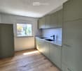 Modern kitchen with light grey cabinets, white countertops, and a double sink. The room features wooden flooring and a window letting in natural light.