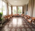 Bright balcony with large windows, wicker chairs with white cushions, patterned tile floor, and potted plant.