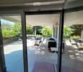 Balcony with tiled floor, glass sliding doors, outdoor furniture including a table, chairs, a grill, and a small stool. The balcony has a view of a garden with trees.