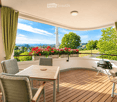 Wooden balcony with a table, chairs, and a grill, overlooking a scenic view with trees and a cityscape.