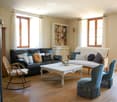 Bright living room with wooden flooring, large windows, a black leather sofa, a white coffee table, a rocking chair, and a blue armchair.