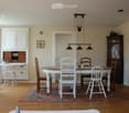 Bright dining area with wooden floor, large wooden dining table surrounded by white and wooden chairs, a hanging light fixture, a white cabinet with drawers, a small desk with a chair, and a dark wooden cabinet with glass doors.