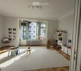 Bright, airy room with wooden herringbone flooring, large white rug, large windows with open shutters, and various children's furniture and toys.