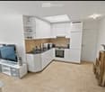 Modern, compact kitchen and living area with white cabinets, a microwave, oven, sink, and a television on a stand. The space includes a small wooden stool and a glass table.