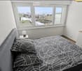 Modern bedroom with large windows offering a city view, featuring a grey upholstered bed with a black and white leaf-patterned duvet, a nightstand with a lamp, and a white cabinet with wooden trim.