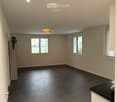 Empty room with light-colored walls, dark wooden flooring, and multiple windows allowing natural light. The room features recessed ceiling lights and a door leading to another area.