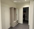 Empty hallway with grey tiled floor, white walls, and a built-in storage unit on the left. A doorway leads to a bathroom on the right.