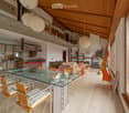 Open-plan living and dining area with a glass dining table, modern chairs, a sofa, kitchen area with counter and stools, and decorative items including a guitar and a flower vase.