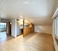 Bright, empty room with wooden flooring and white walls, featuring large windows and a small kitchen area with cabinets and appliances visible in the background.
