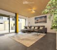 Modern living room with a gray sofa, coffee table, and a beige rug. The room features a large glass door leading to a patio, a picture frame on the wall, and a potted plant.